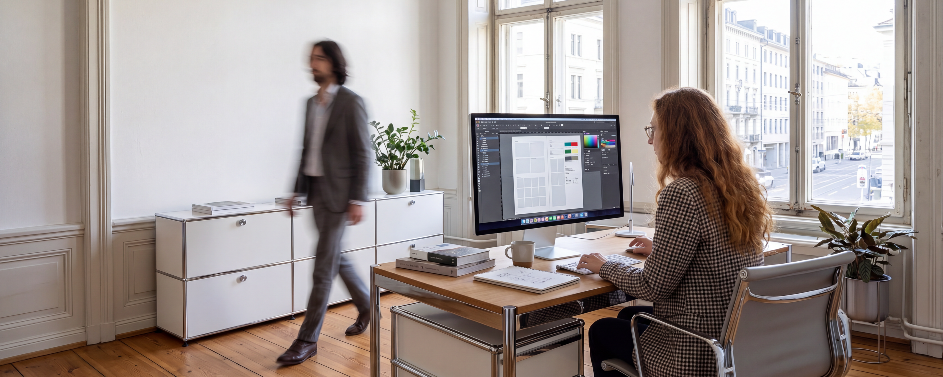we-dot-Büro im Altbau: eine Mitarbeiterin gestaltet ein Layout am iMac, ein Kollege geht im Hintergrund vorbei