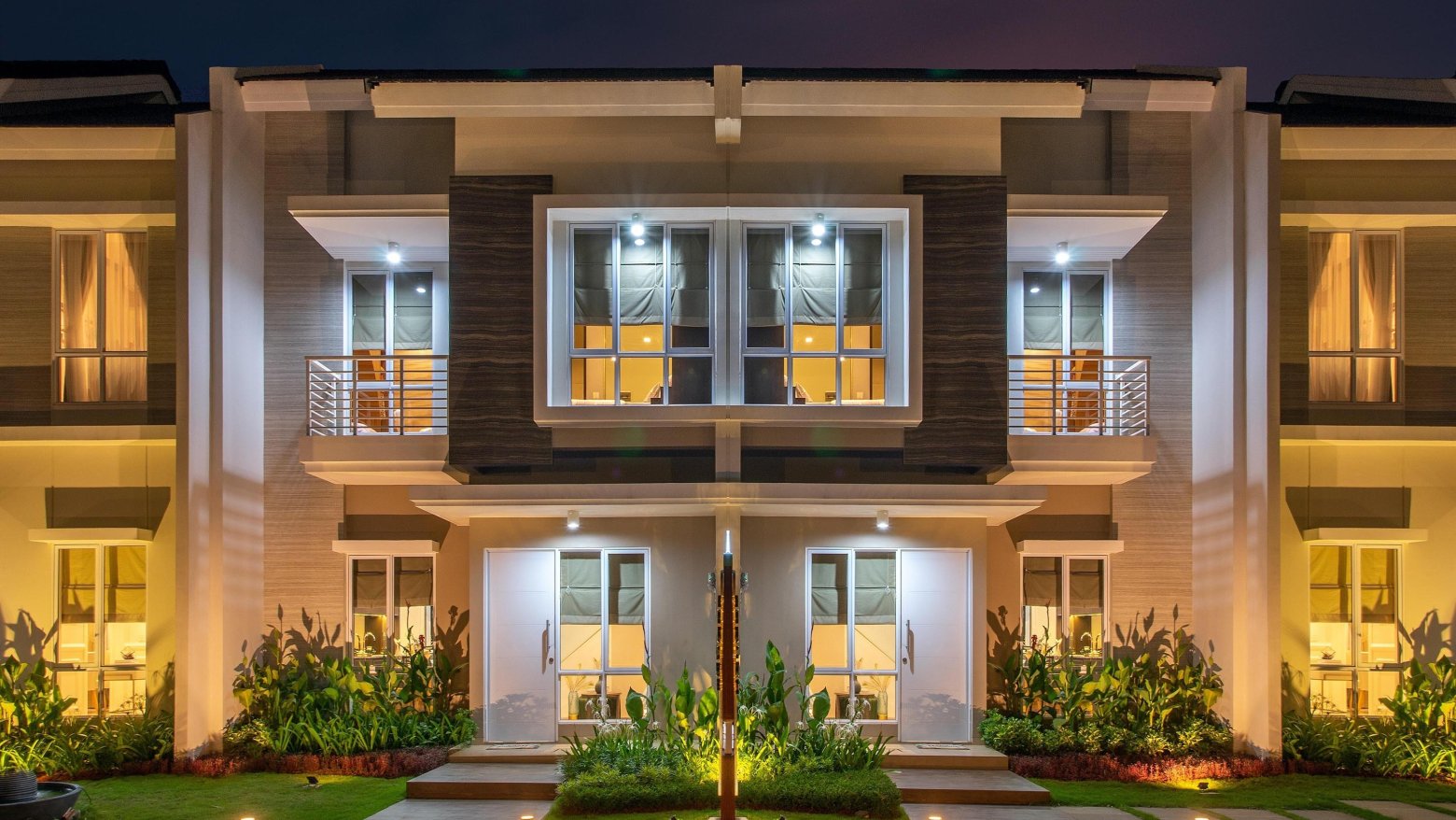 Elegante, beleuchtete Doppelhaushälfte mit modernen Balkonen und gepflegter Gartenlandschaft, ideal für Immobilienmarketing.